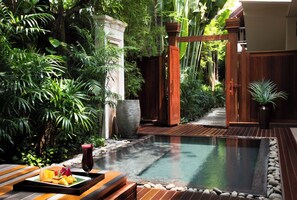 Un área de piscina tranquila rodeada de exuberante vegetación, una terraza de madera y un sendero de piedra que lleva a un portón de madera.