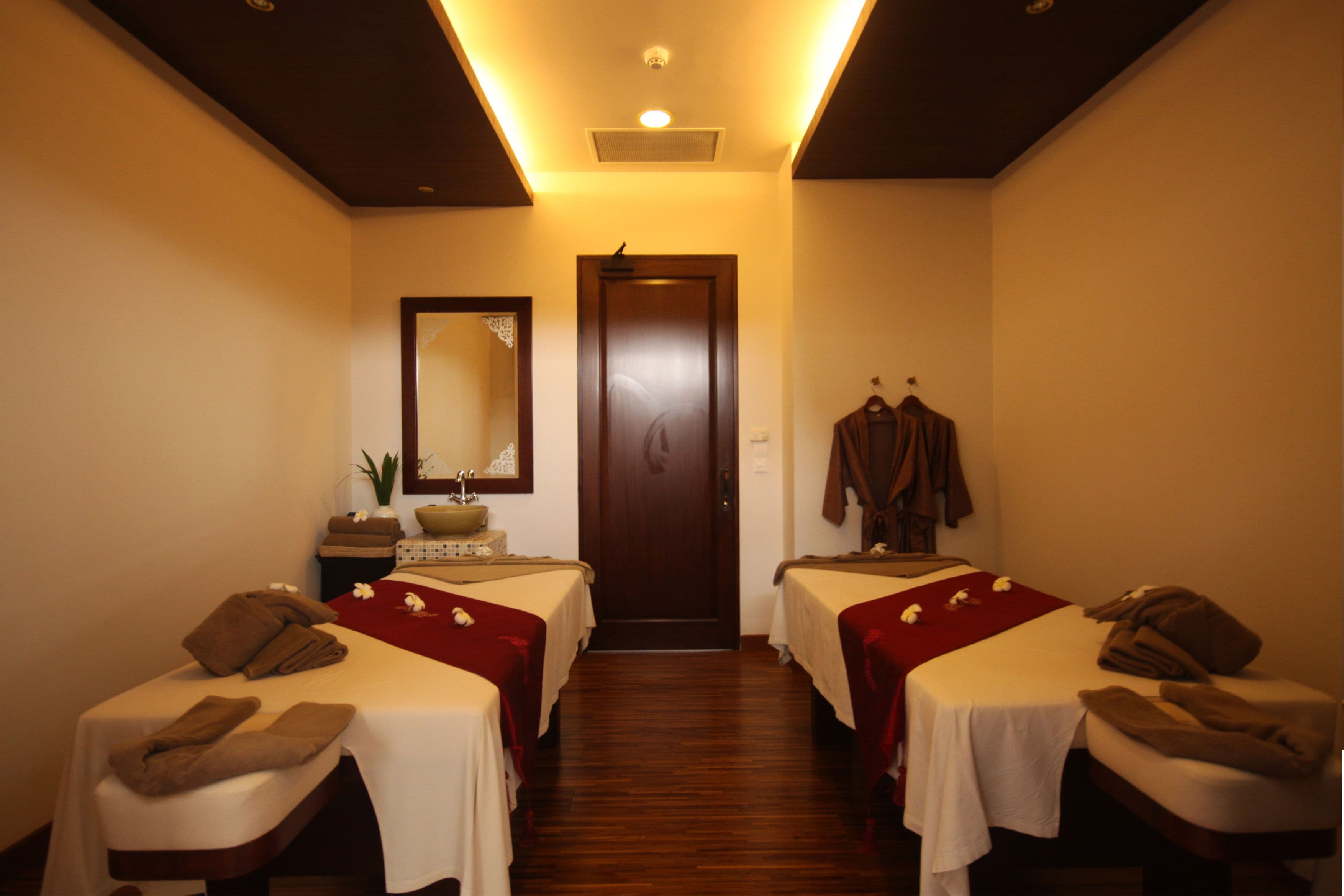 A room with two beds, each with a towel and a small table with a bowl and a plant. There is a mirror on the wall and a door at the end of the room.