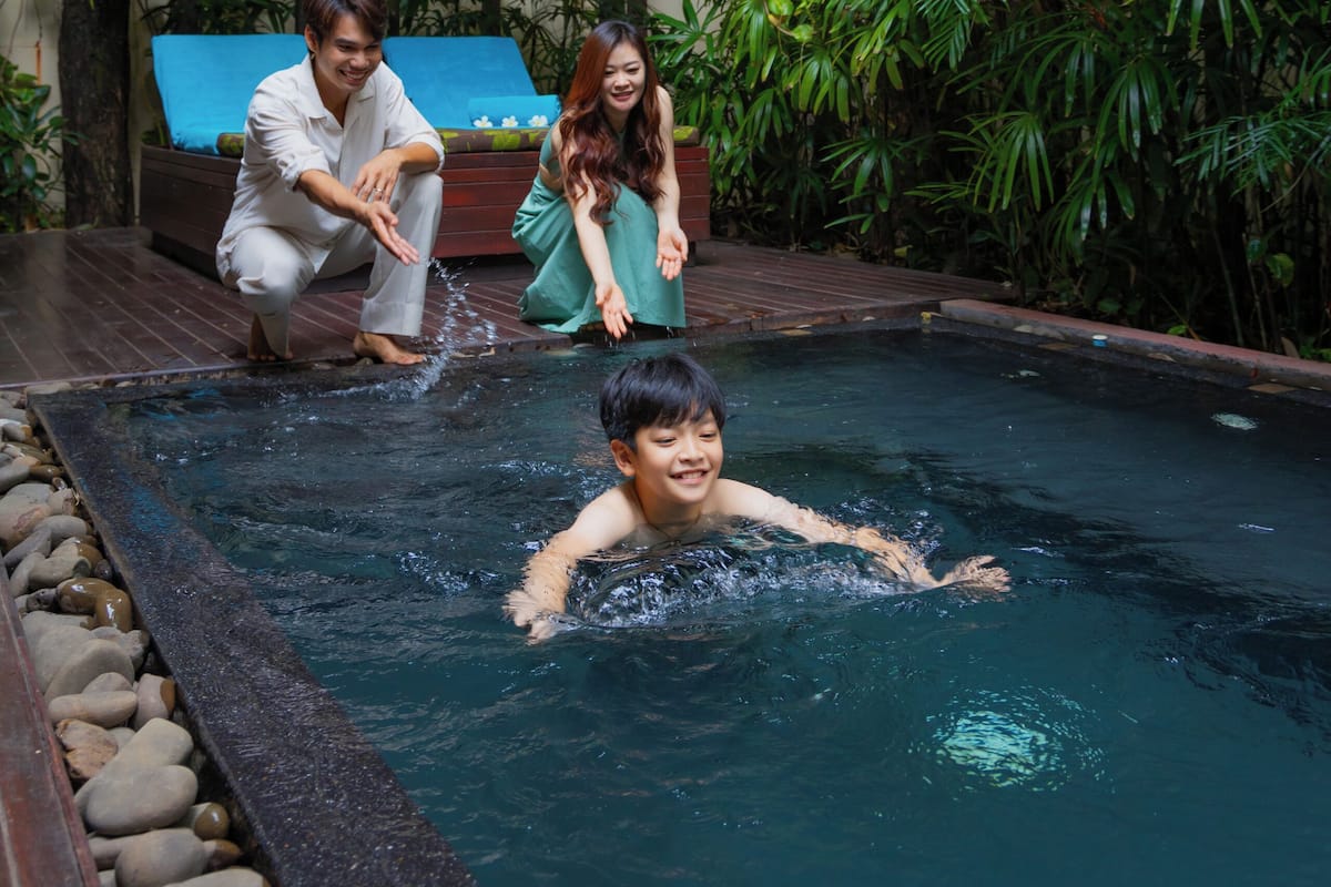 A pool area with a wooden deck, a lounge chair, and lush greenery.