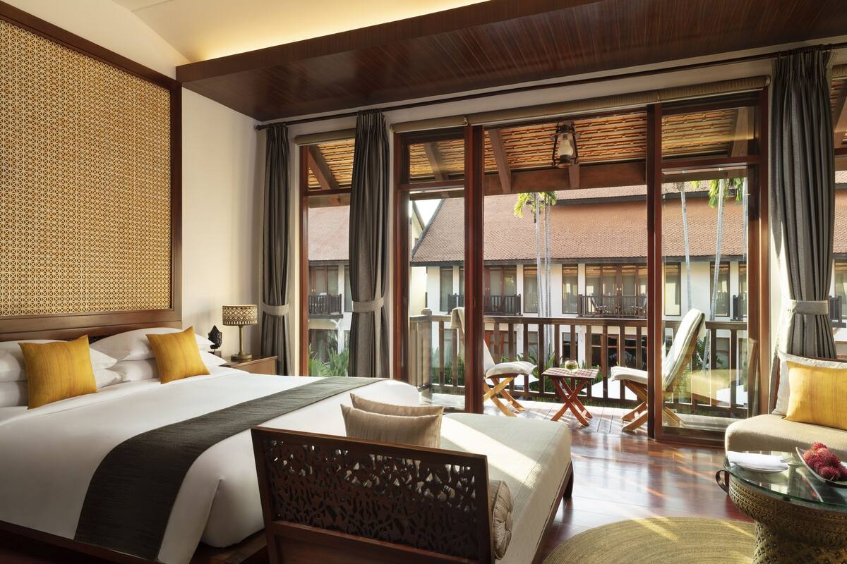 A hotel room with a large bed, a sitting area with a glass table, and a balcony with outdoor furniture.