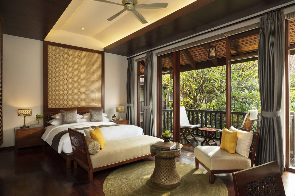 A spacious bedroom with a large bed, a ceiling fan, a view of greenery outside, and a balcony with seating.