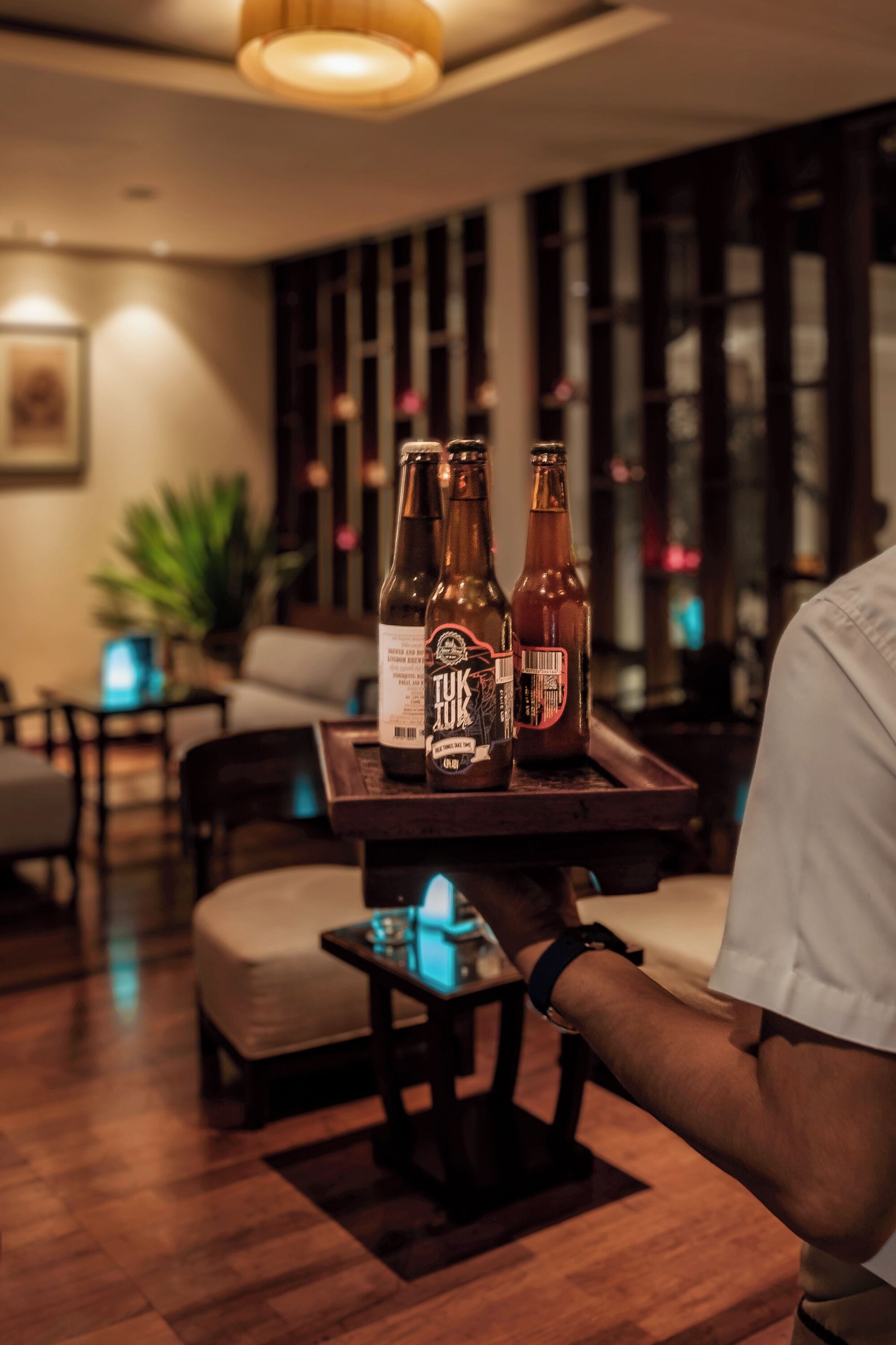 Una zona de bar con muebles de madera, una bandeja con dos botellas de cerveza y una persona con camisa blanca.