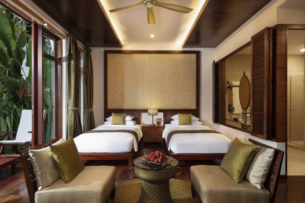 A hotel room with two beds, a sitting area with a coffee table, a ceiling fan, and a large window with curtains.