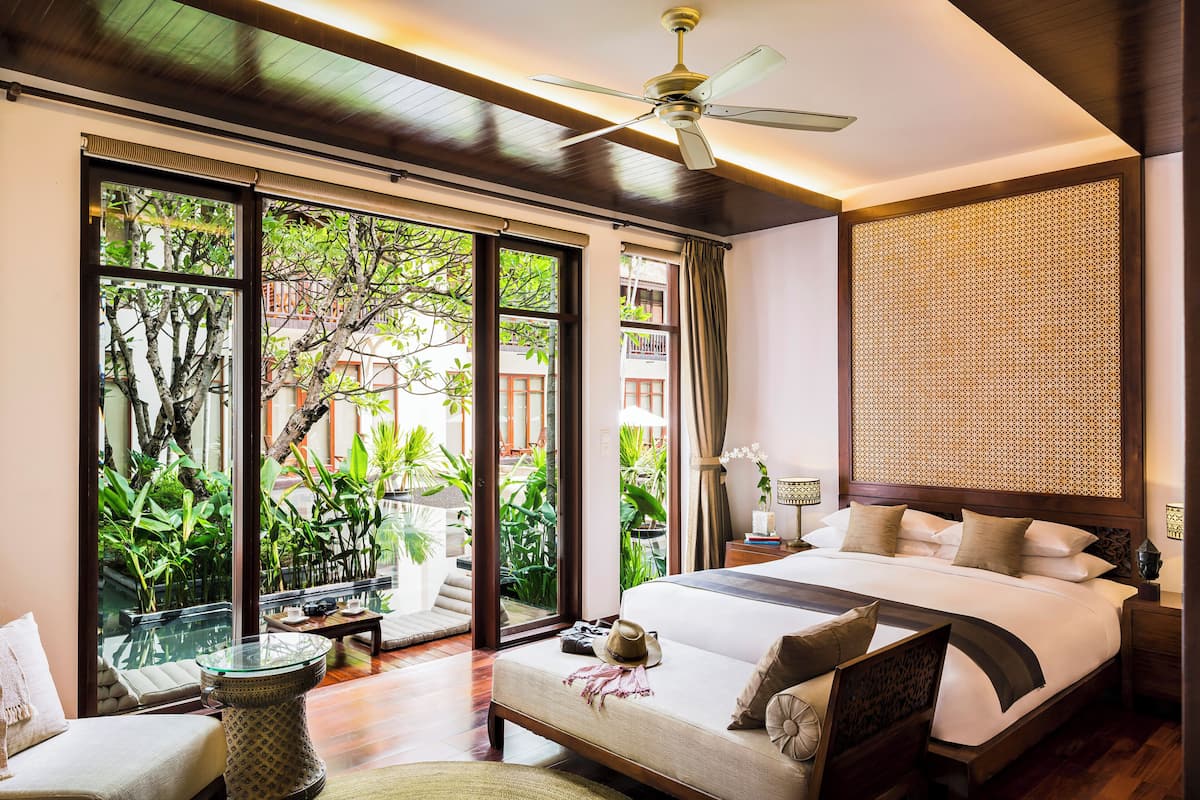 A spacious bedroom with a large bed, a ceiling fan, and a view of lush greenery outside.