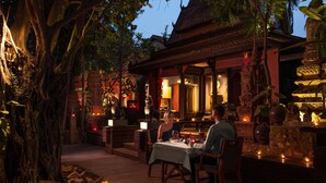 An outdoor dining area with a table set for two, surrounded by decorative elements and a large tree. - Anantara Angkor Resort (Siem Reap)