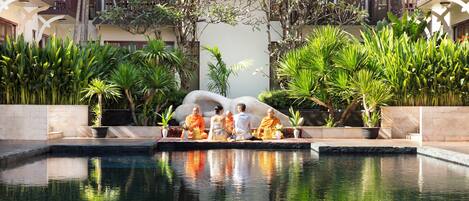 A serene pool area with a statue of monks, surrounded by lush greenery and a multi-story building with balconies.