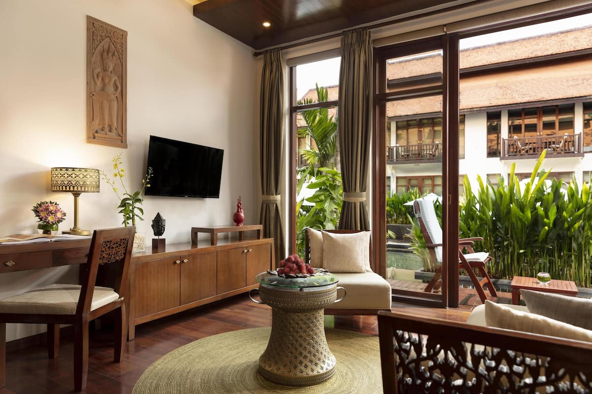 A well-lit room with a flat-screen TV, wooden furniture, a decorative table, and a view of a balcony with plants and a pool.