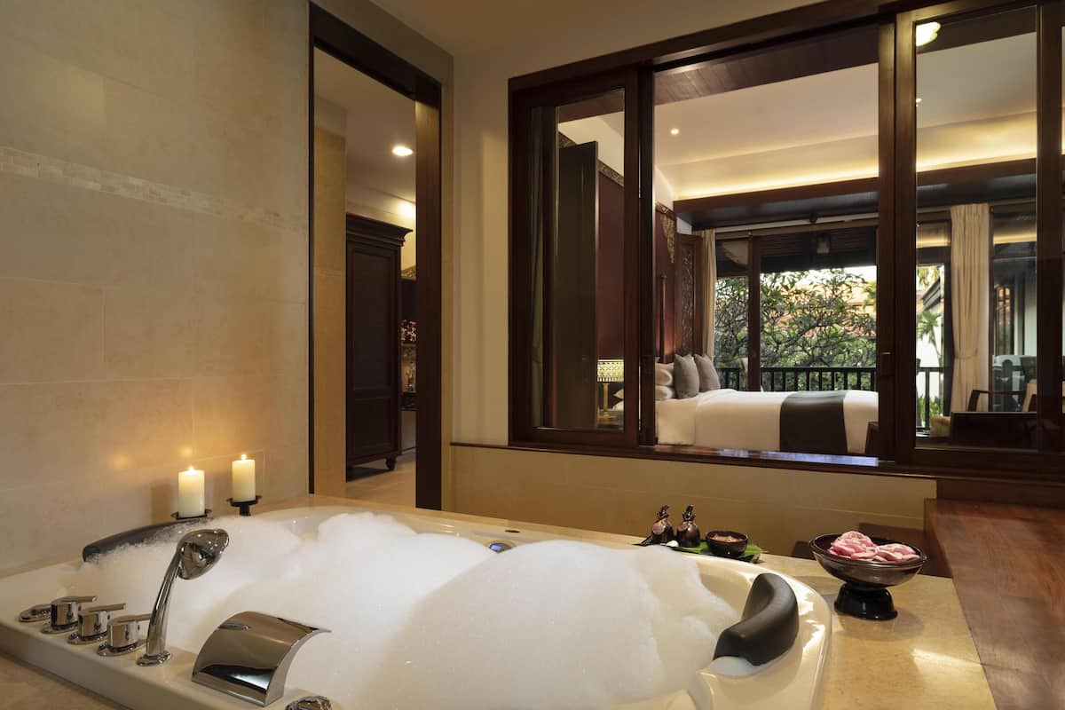 A luxurious bathroom with a large bathtub, candles, and a mirror reflecting a bedroom with a balcony.