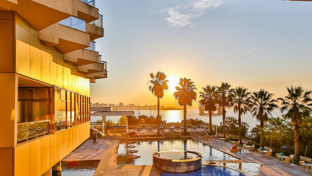 A modern building with a pool area, including a circular hot tub and a rectangular pool, surrounded by palm trees and a sunset view. - Quality Niteroi (NITEROI)