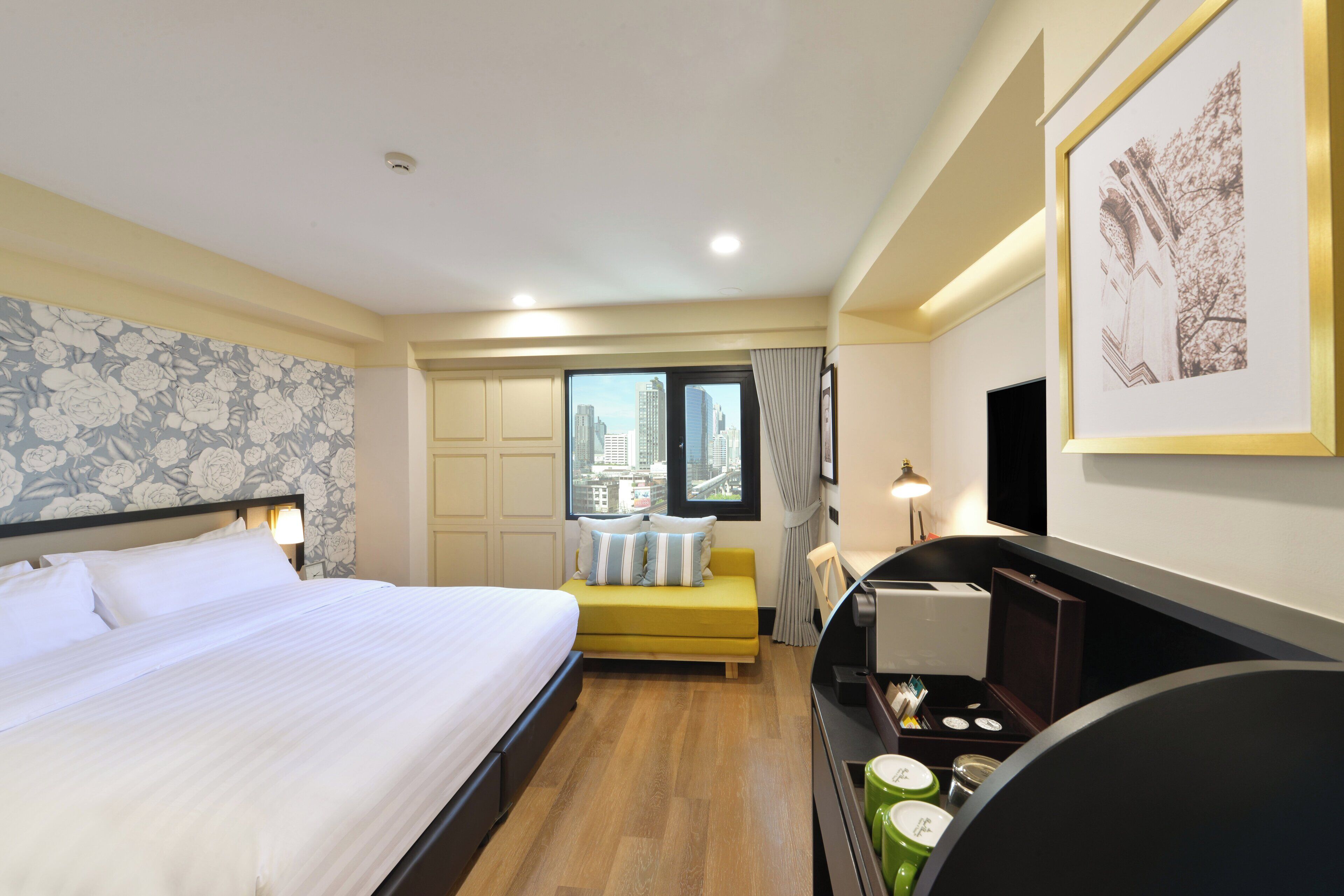 A hotel room with a large bed, a yellow sofa, a desk with a lamp, and a city view through the window.