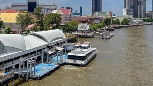 A waterfront area with a modern high-rise building, a covered dock with boats, and a cityscape in the background. - Oakwood Hotel & Residence Bangkok (Bangkok)