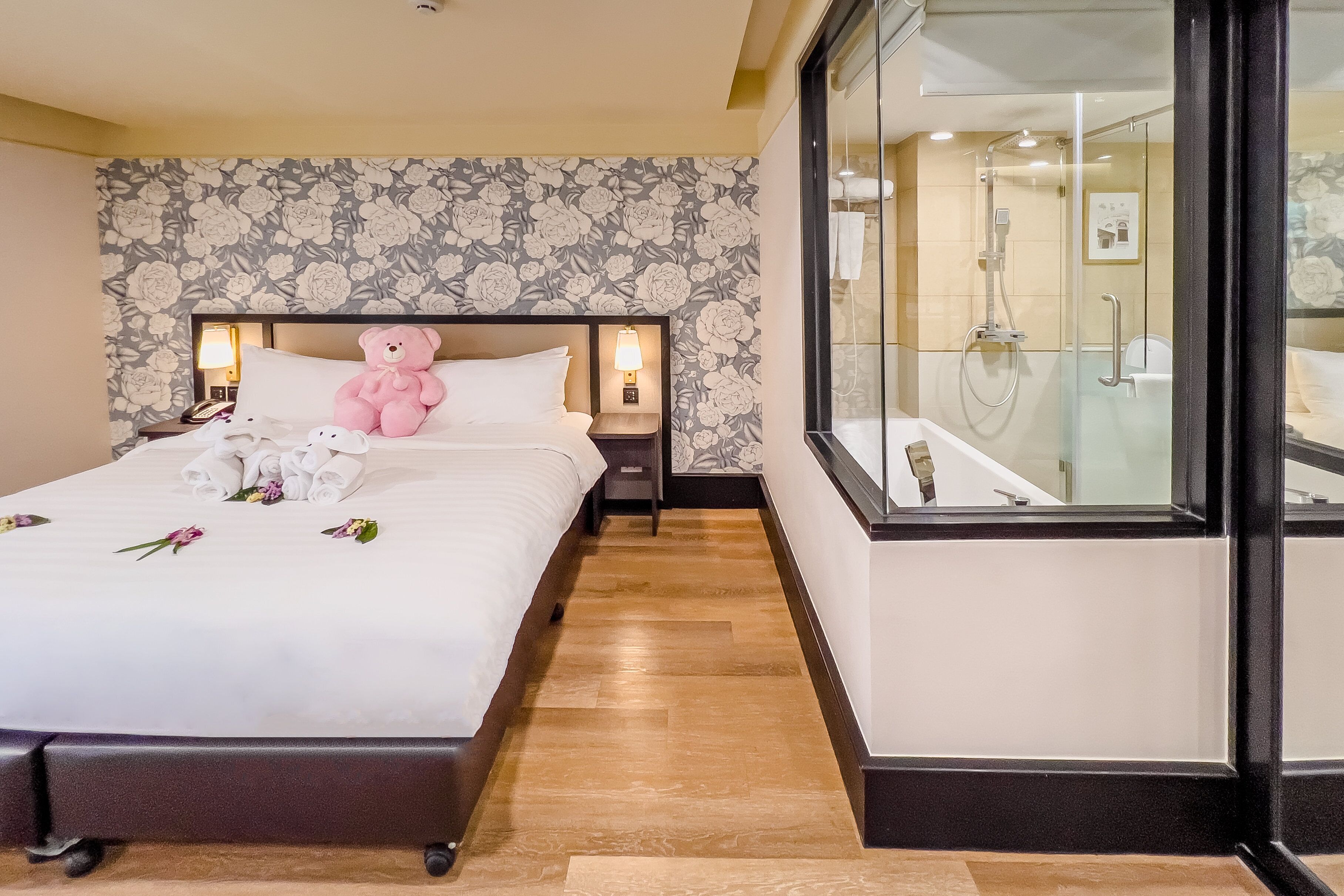 A neatly made bed with a teddy bear, a nightstand with a lamp, and a view of a glass-enclosed shower area.