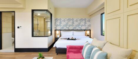 Una habitación de hotel moderna con una cama grande, un sofá, una mesita con una planta y un baño visible a través de una puerta de cristal.