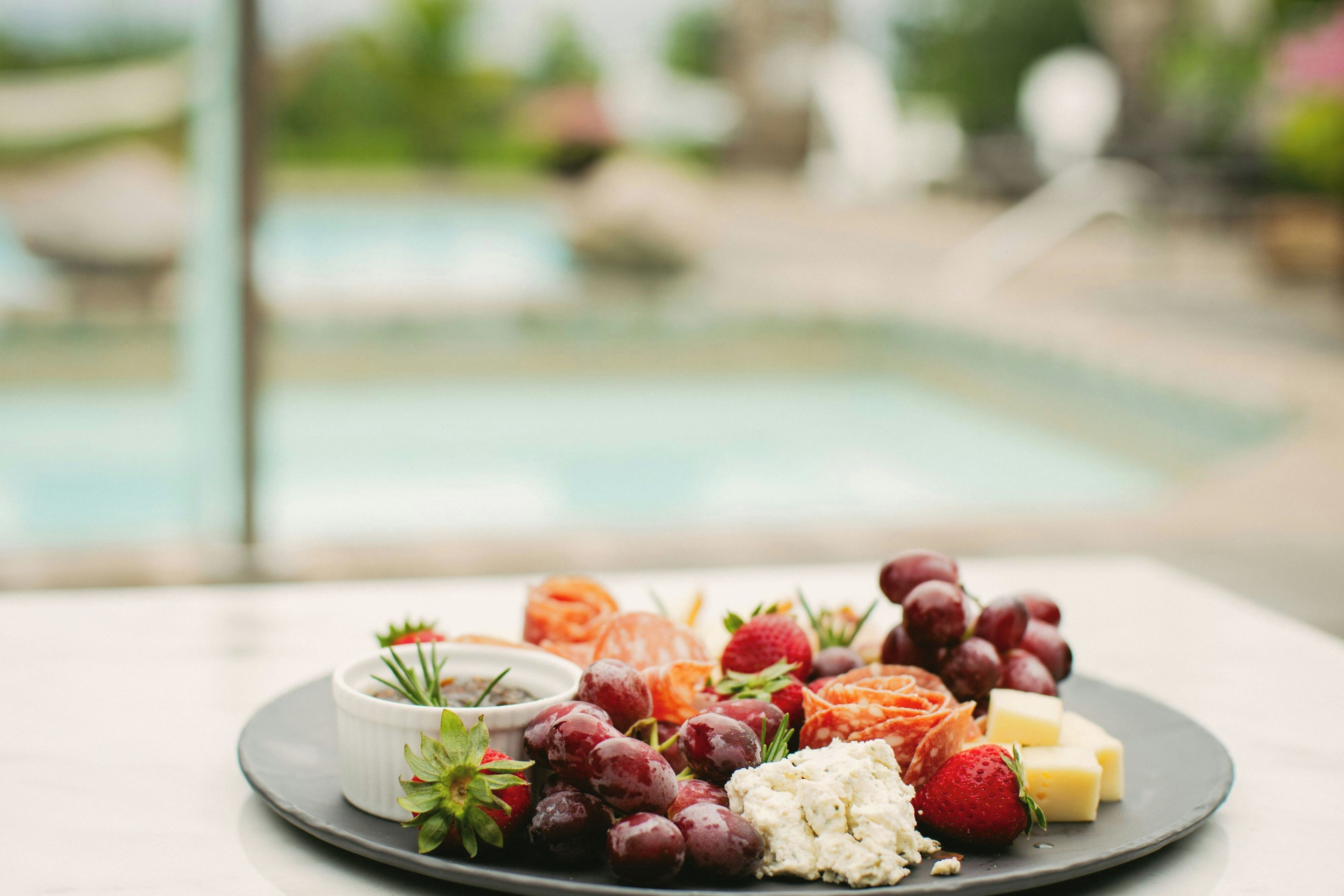 Un plato con quesos variados, uvas y fresas sobre una mesa, con una piscina al fondo.