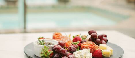Un plato con quesos variados, uvas y fresas sobre una mesa, con una piscina al fondo.