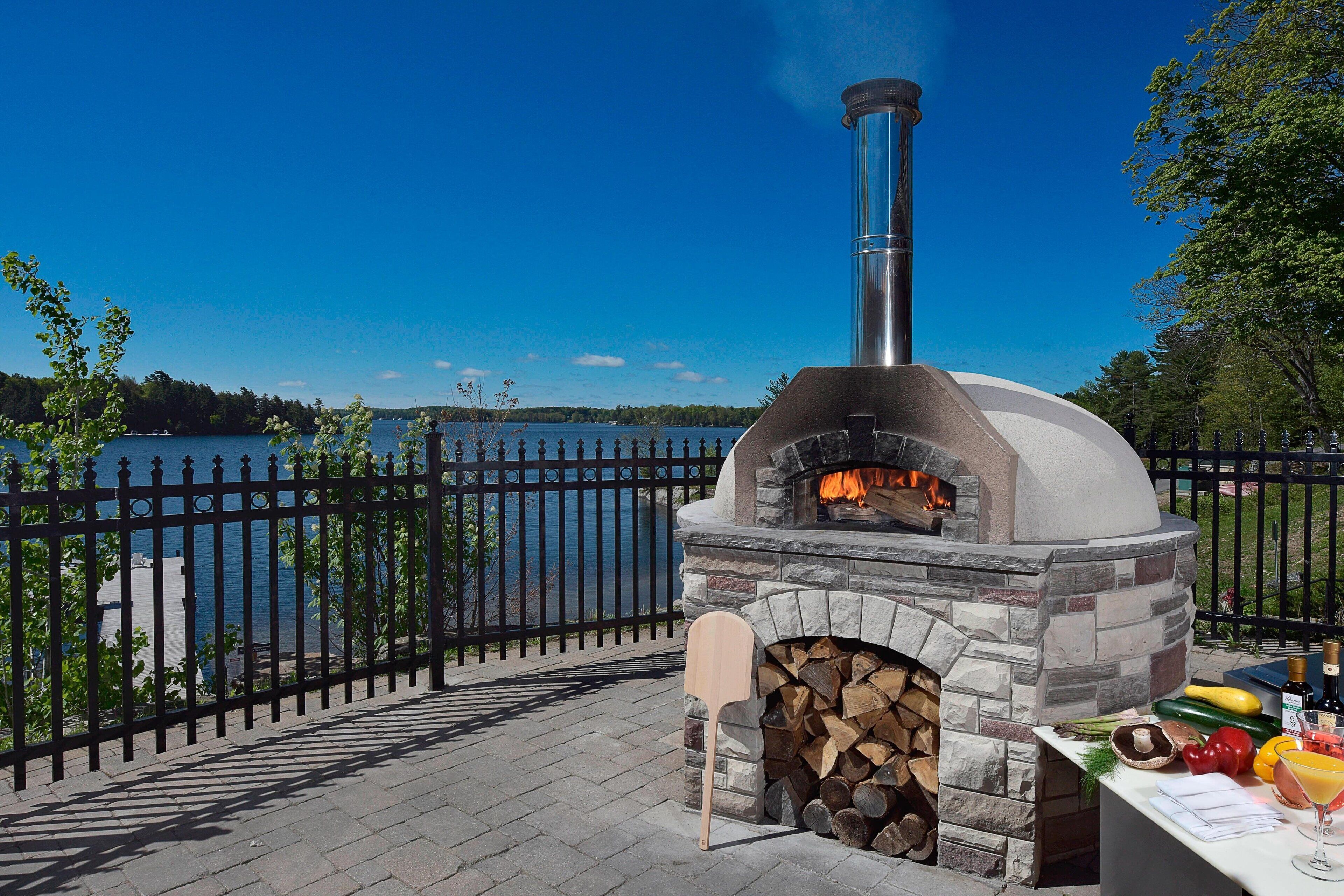 Un horno de pizza al aire libre con fachada de piedra y chimenea, situado junto a un lago con una cerca de metal negro.