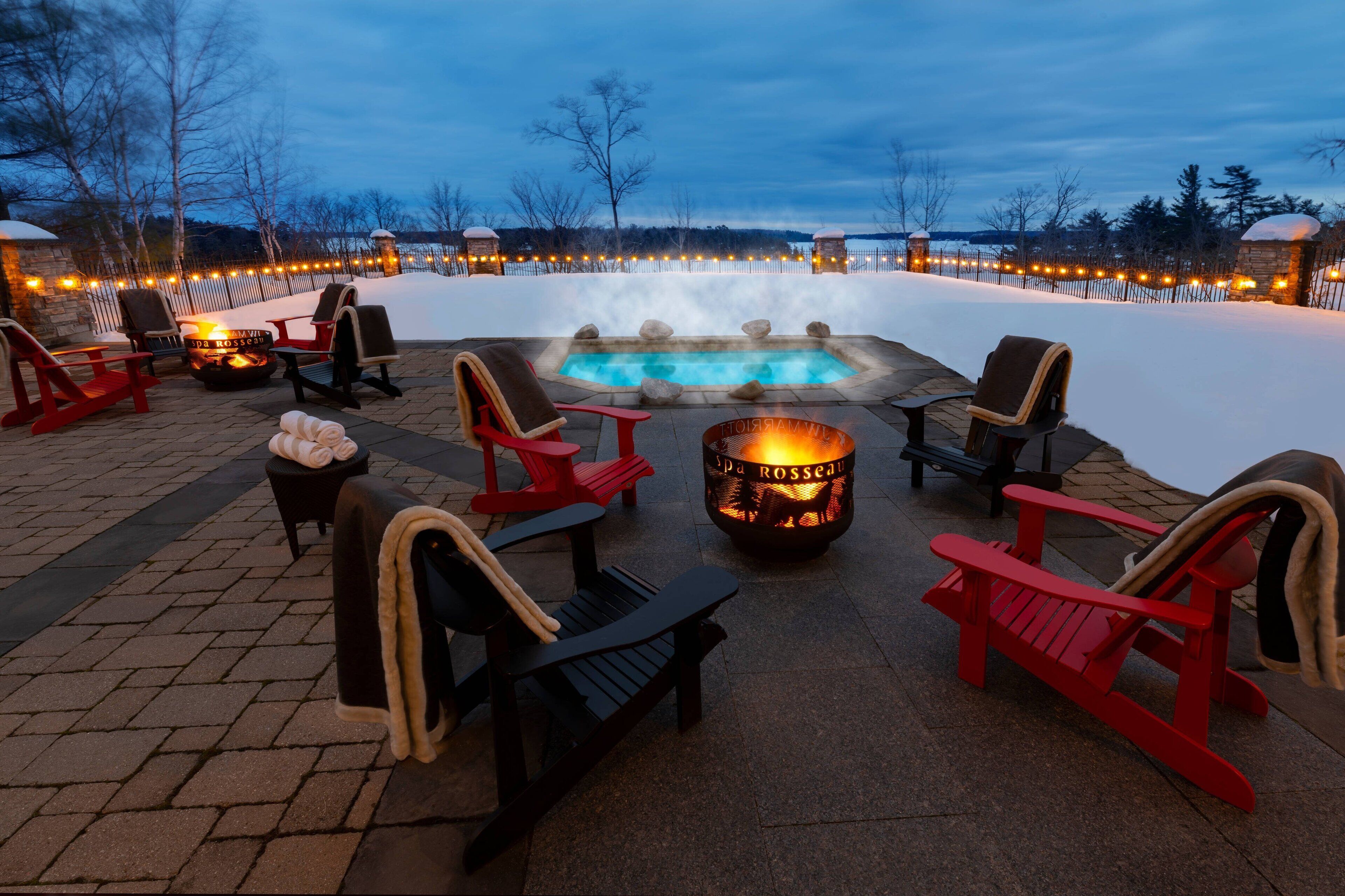 A heated outdoor seating area with Adirondack chairs, a fire pit, and a hot tub surrounded by a snow-covered landscape.