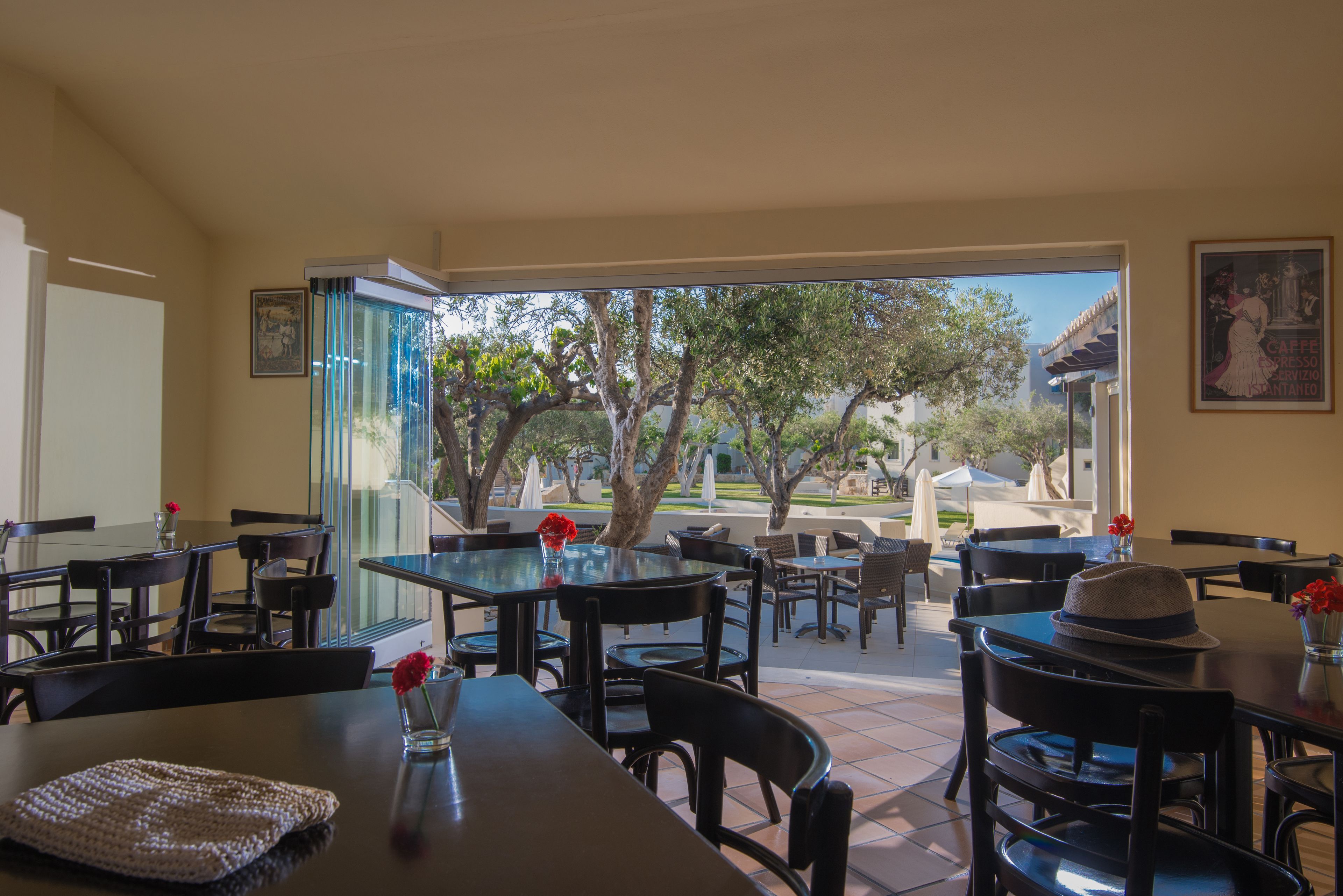 An outdoor dining area with tables and chairs, a view of trees and a building in the background, and a framed picture on the wall.