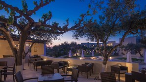 An outdoor patio with wicker furniture and tables, surrounded by trees and a pool area. - Ourania Apartments (Hersonissos)