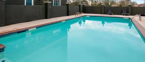 A rectangular swimming pool with a clear blue water surface, surrounded by a brick deck and a low stone wall. There are two lounge chairs and a small window on the wall.