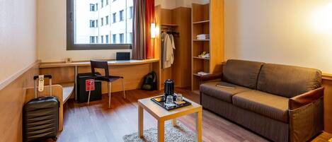 A compact hotel room with a sofa, a small table, a desk with a laptop, a chair, a suitcase, and a wardrobe.
