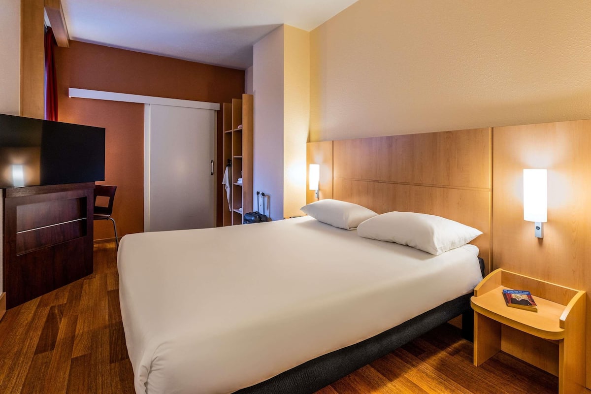 Habitación de hotel con cama, mesita de noche, televisión, armario y espejo.