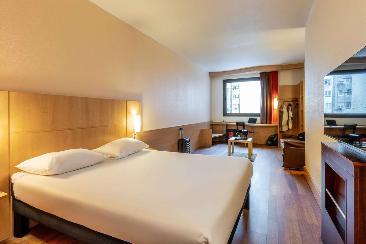 A hotel room with a large bed, a desk, a chair, a laptop, and a window with curtains.