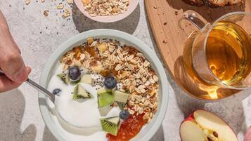 Eine Schüssel Joghurt mit Früchten und Granola, ein Glas bernsteinfarbene Flüssigkeit und Scheiben Brot auf einem Tisch.