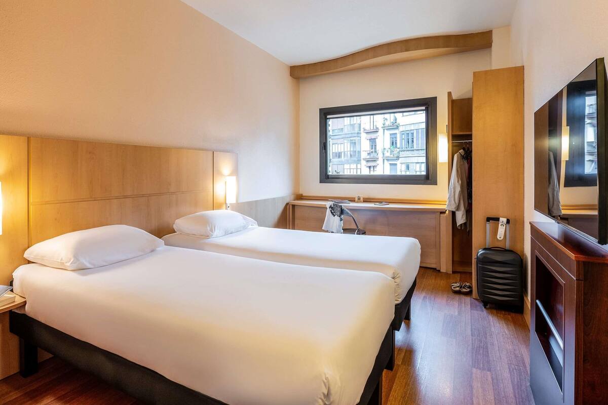 A hotel room with two beds, a desk, a mirror, a TV, and a window with a city view.