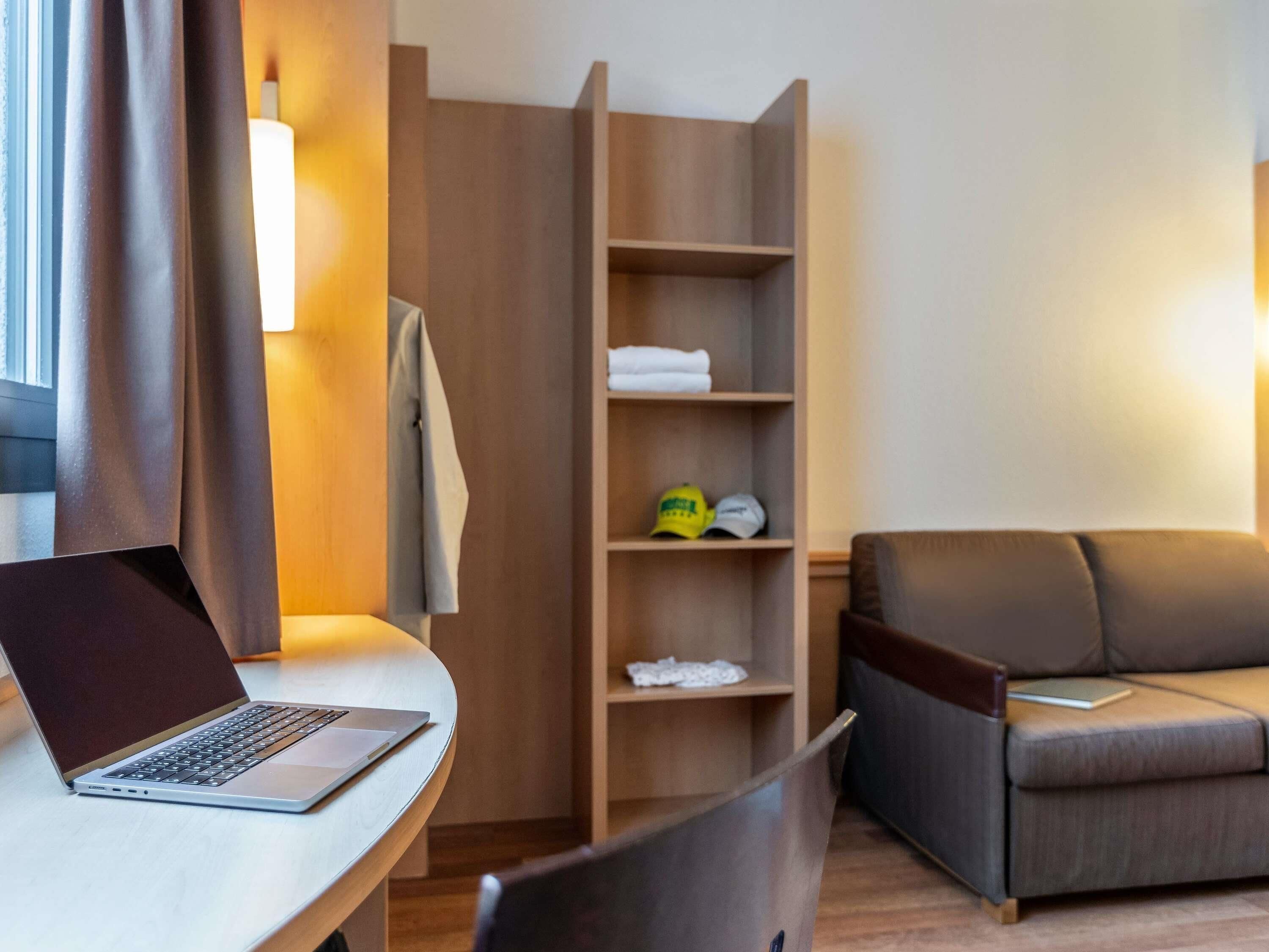 Una habitación de hotel moderna con una laptop sobre un escritorio, un sofá y un armario de madera con estantes.