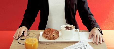 Una persona sentada a una mesa con un croissant, una taza de café y un vaso de zumo de naranja. También se ve un periódico sobre la mesa.