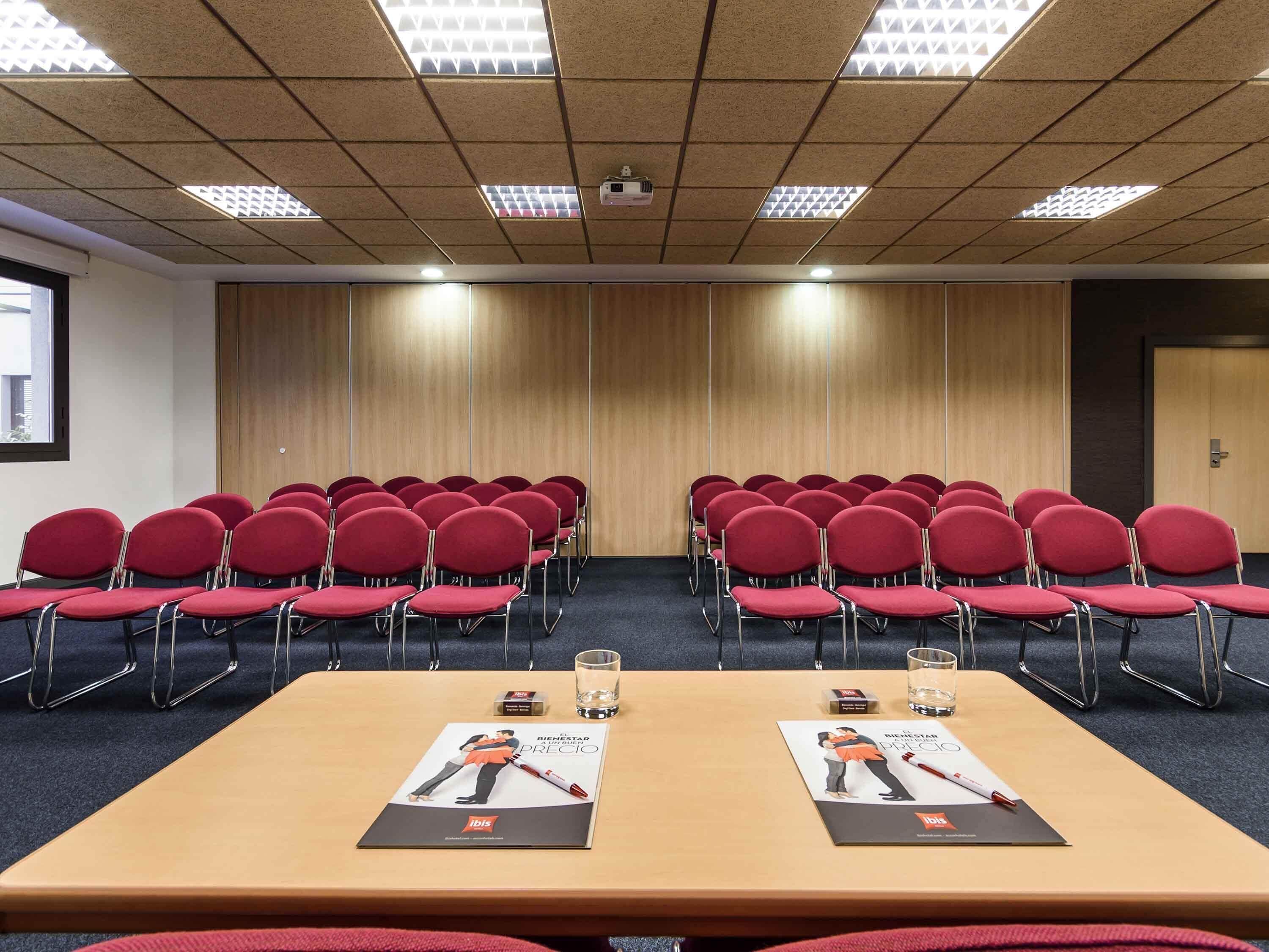 Un salón de conferencias con filas de sillas rojas, un panel de madera en la pared y un techo con luces fluorescentes.
