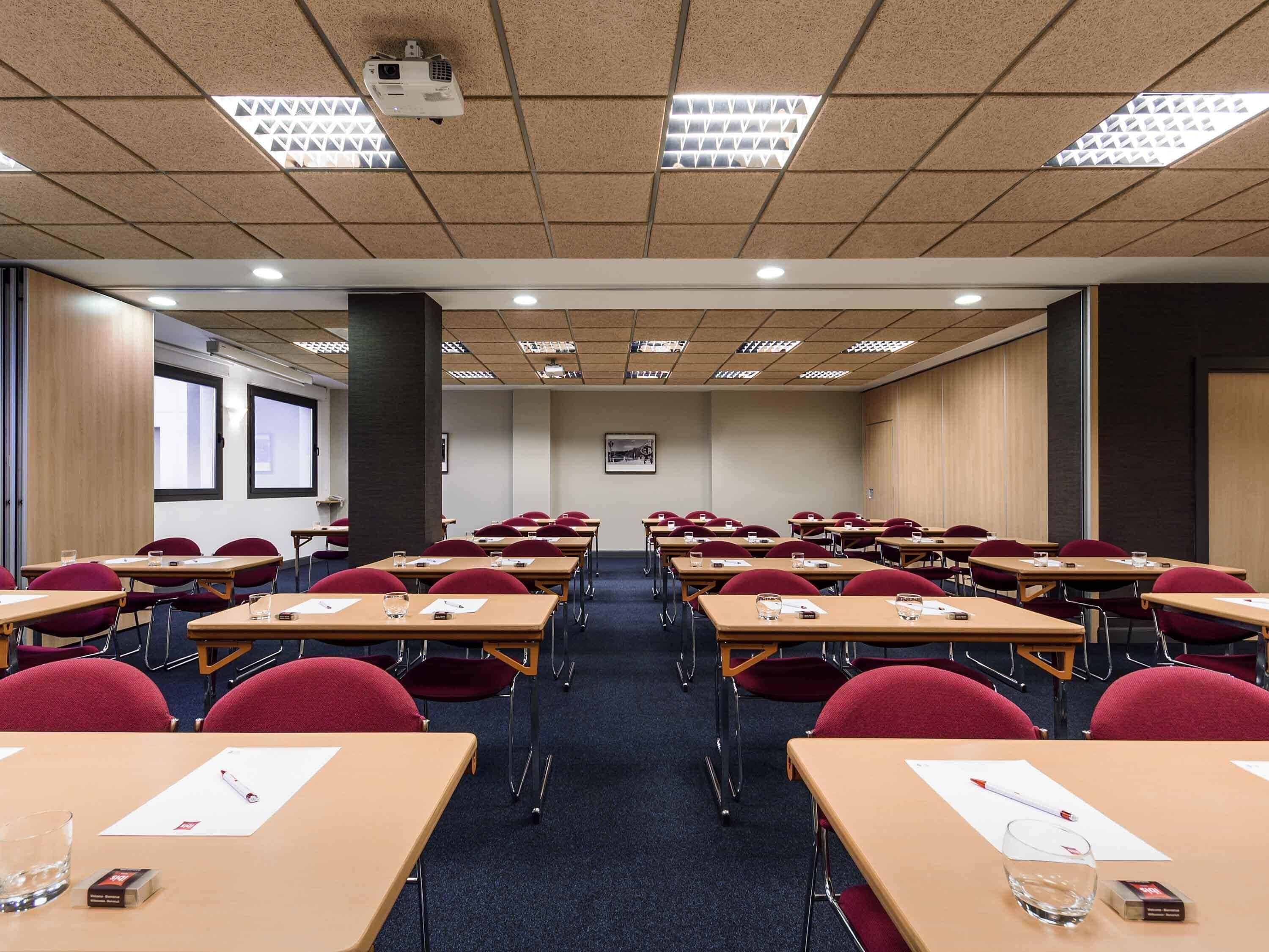 Un salón de conferencias con filas de mesas, sillas y un proyector en el techo.