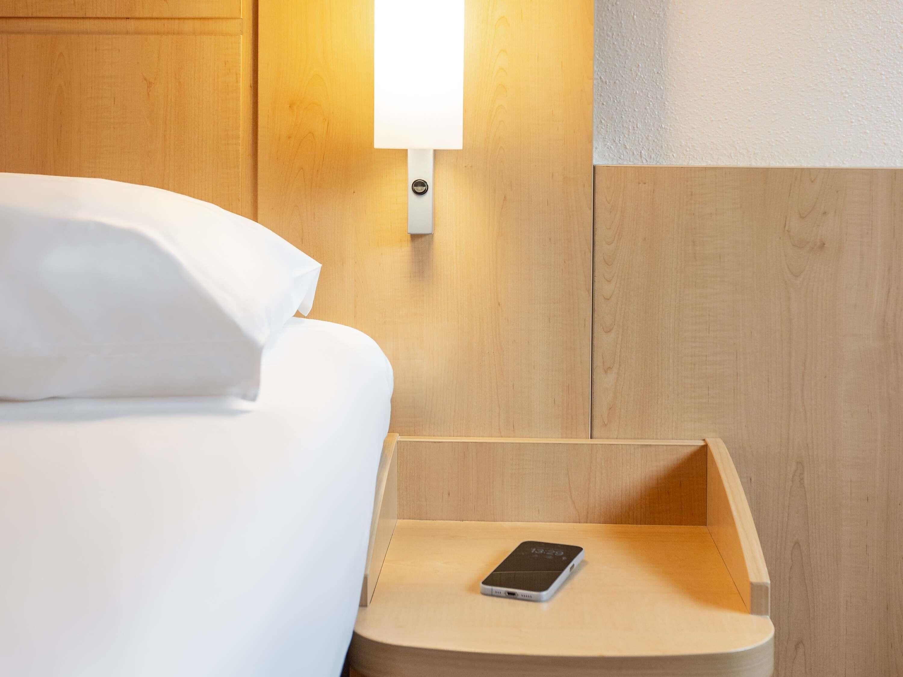Un lit bien fait, avec du linge de lit blanc, et un téléphone intelligent posé sur une table de chevet en bois.