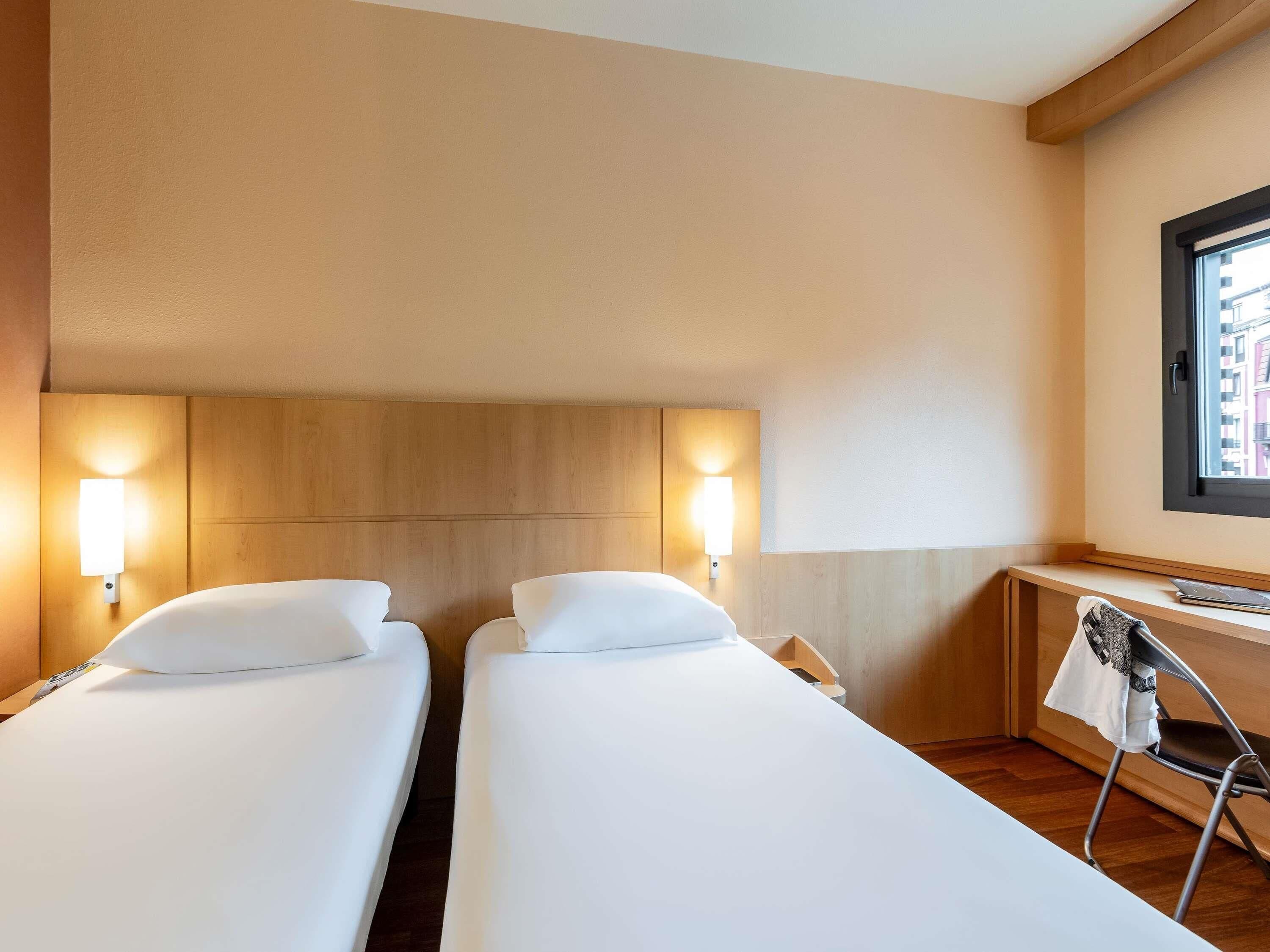 A hotel room with two single beds, a wooden headboard, a desk with a laptop, and a window with a view of buildings.