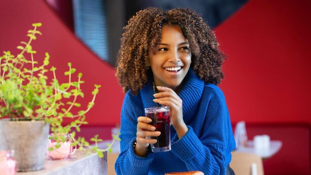 Una persona sentada a una mesa con un sándwich y una bebida, con una planta y una pared roja al fondo.