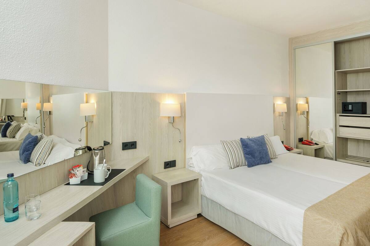 A modern hotel room with a bed, bedside tables, a desk, a chair, a mirror, and a wardrobe.
