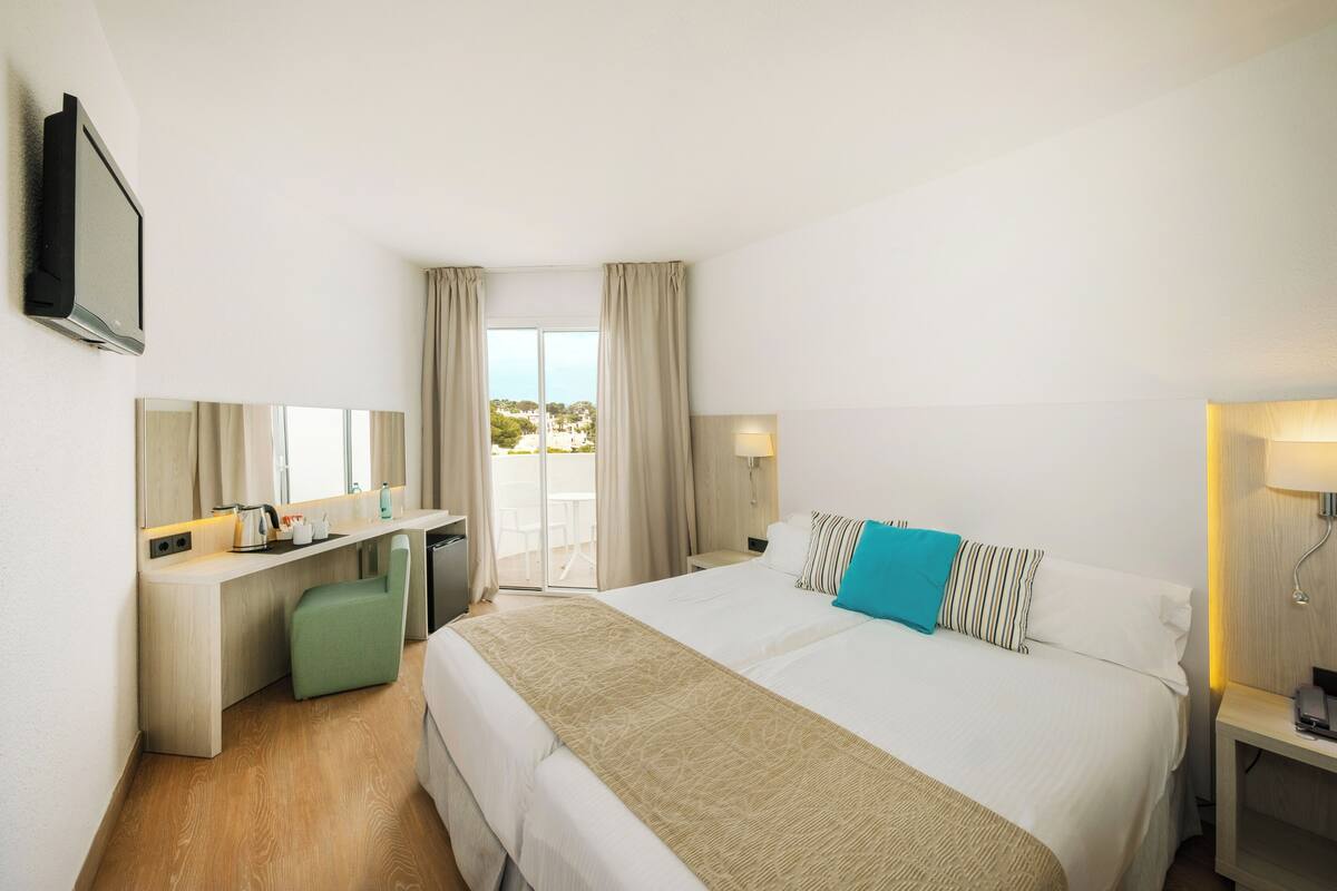 A hotel room with a large bed, a desk, a chair, a TV, and a balcony with curtains.