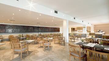 Un vaste espace dédié à la salle à manger, avec des tables et des chaises en bois, une grande fresque murale et, au fond, une cuisine moderne.