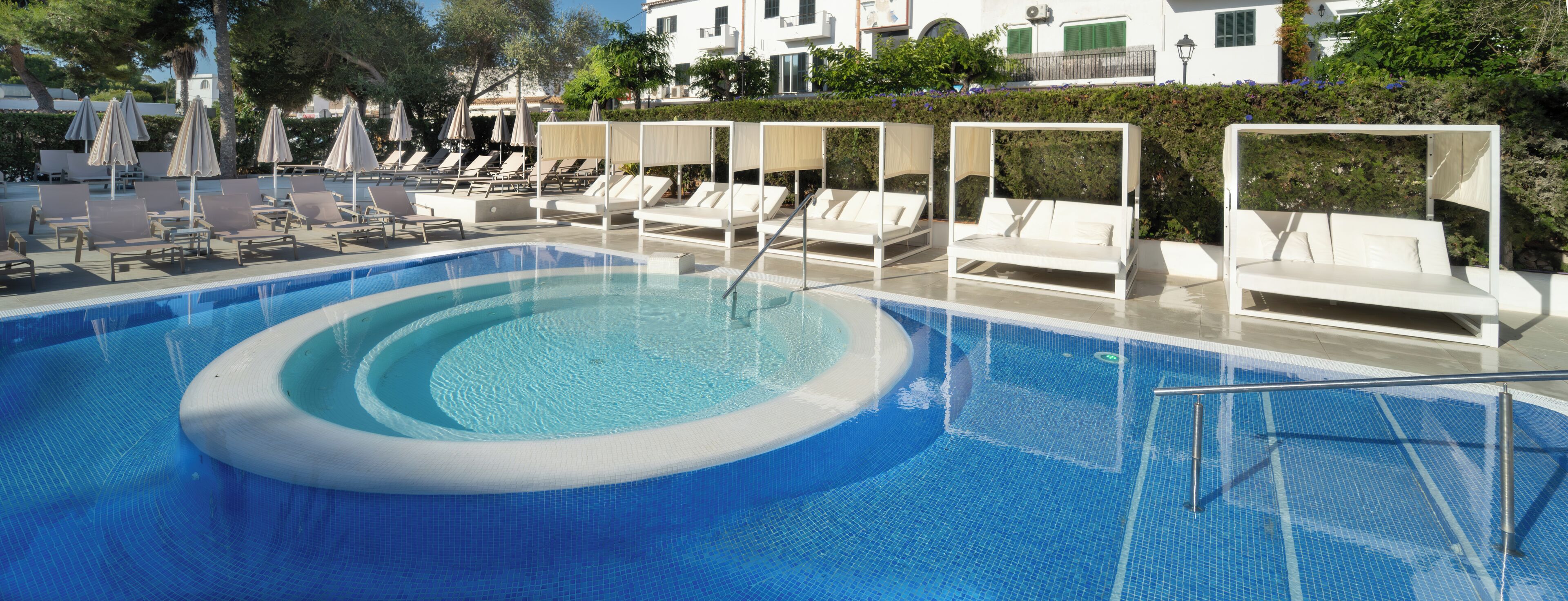 Piscina con sillas de descanso y sombrillas, rodeada de follaje y edificios.