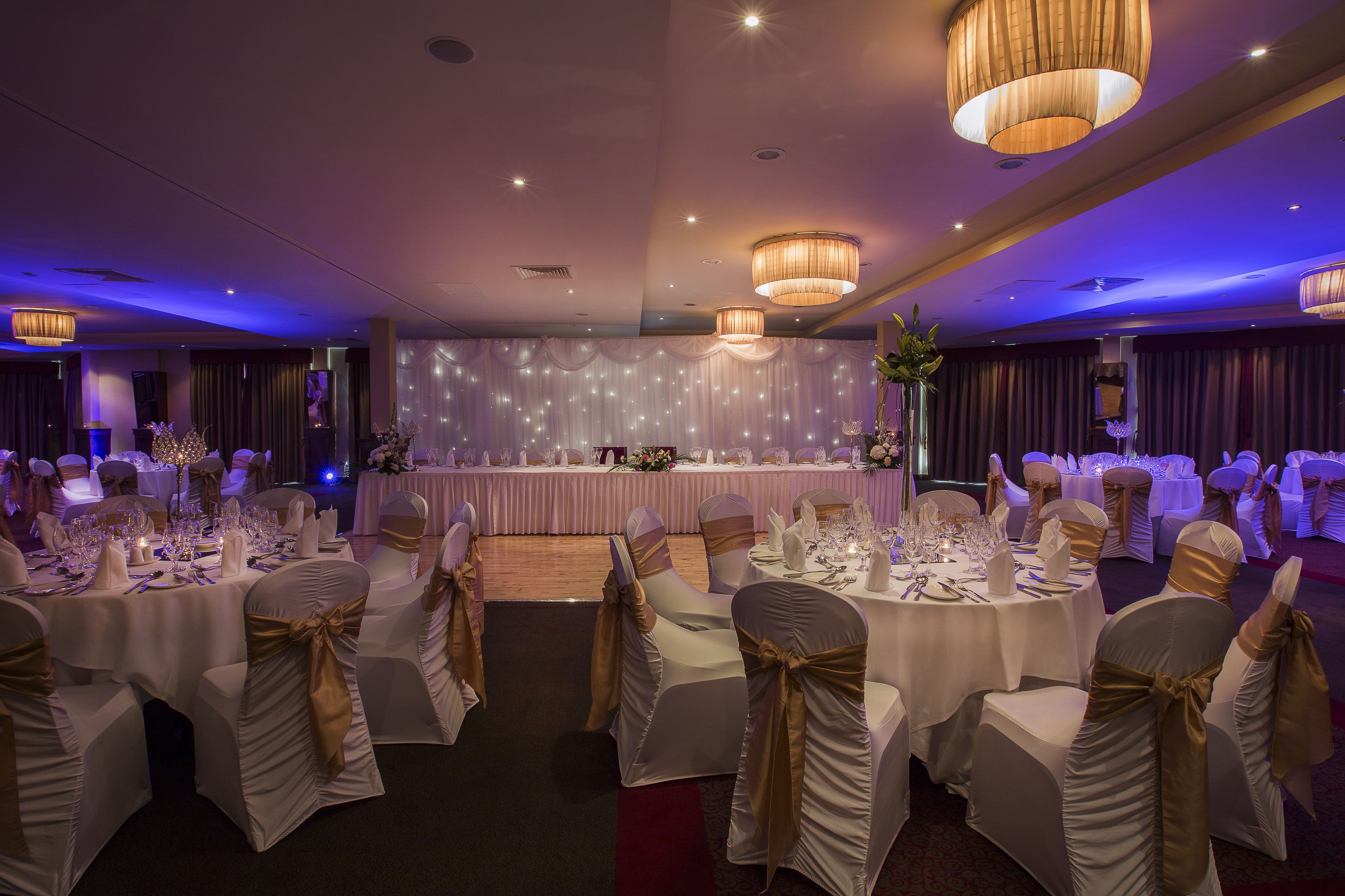 Un salón de banquetes con mesas redondas dispuestas para un evento formal, con manteles blancos y cintas doradas en las sillas.