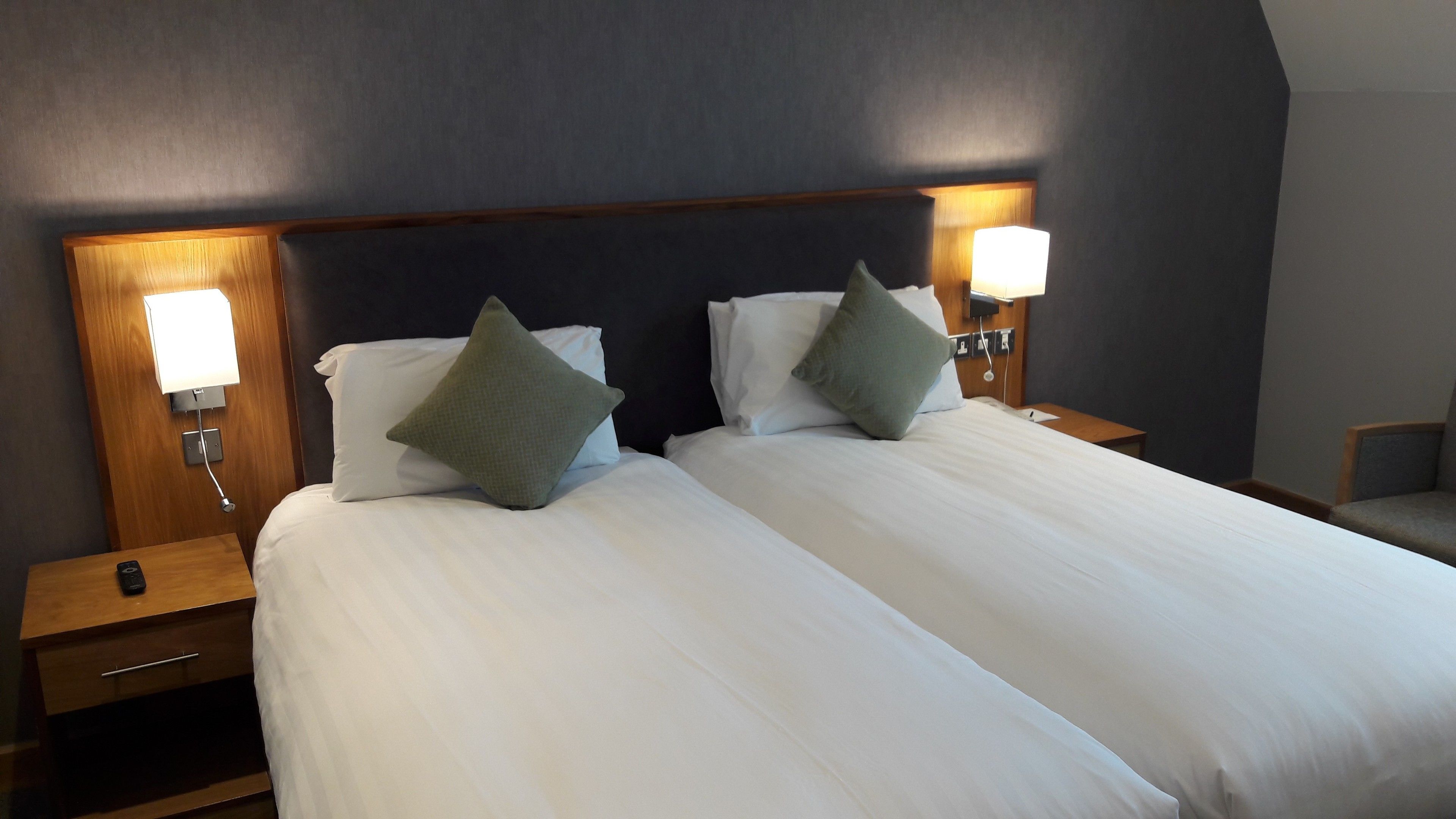 A hotel room with two beds, each with white linens and green pillows. There are bedside tables with lamps and a remote control on one table.