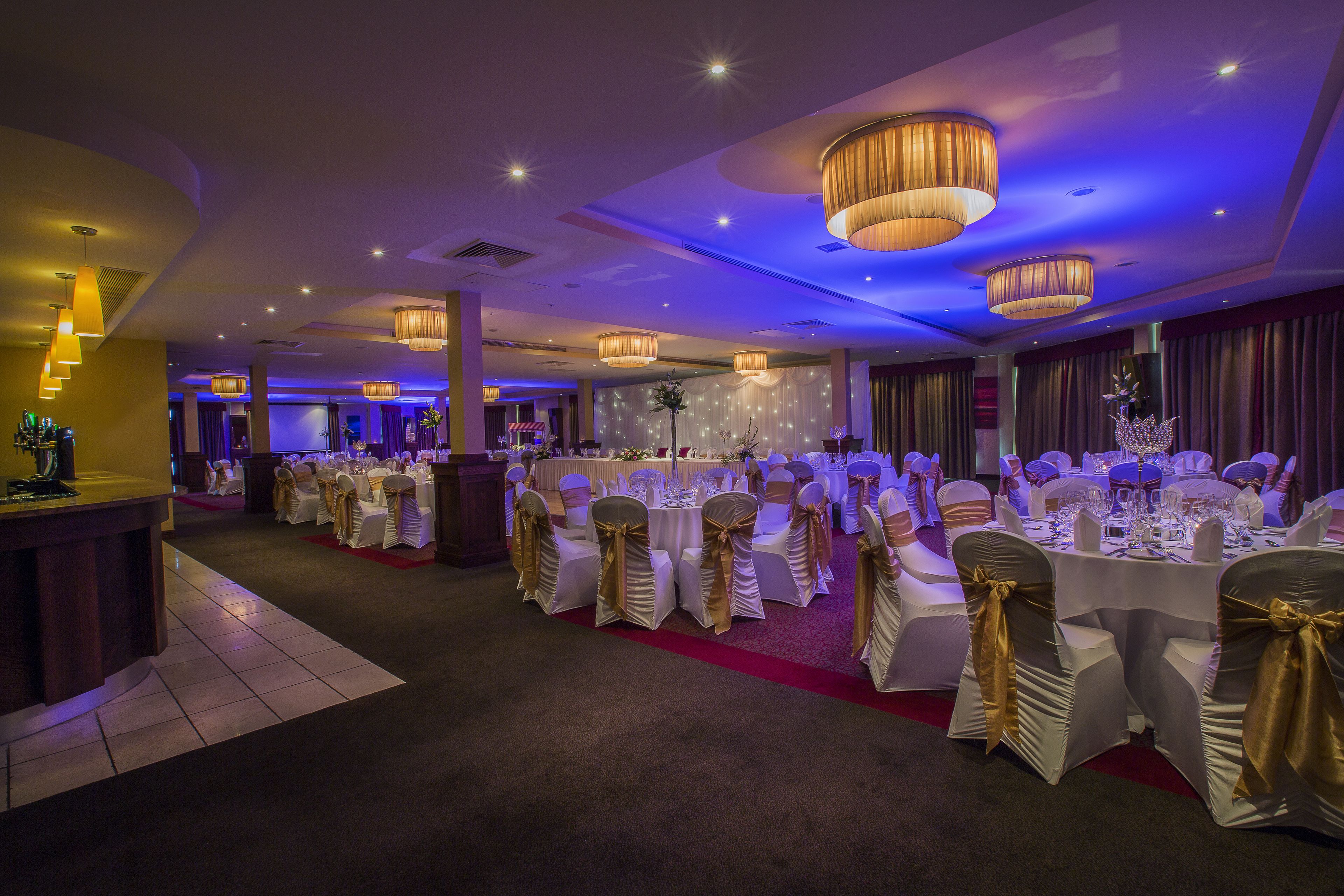 Un salón de banquetes con mesas redondas, manteles blancos y sillas con cintas doradas.