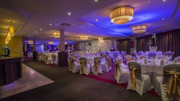 Un salón de banquetes con mesas redondas, manteles blancos y sillas con cintas doradas.