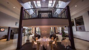 Um hall espaçoso com uma mesa redonda centralmente decorada para um evento formal, uma varanda com grade de ferro forjado e um lustre moderno.
