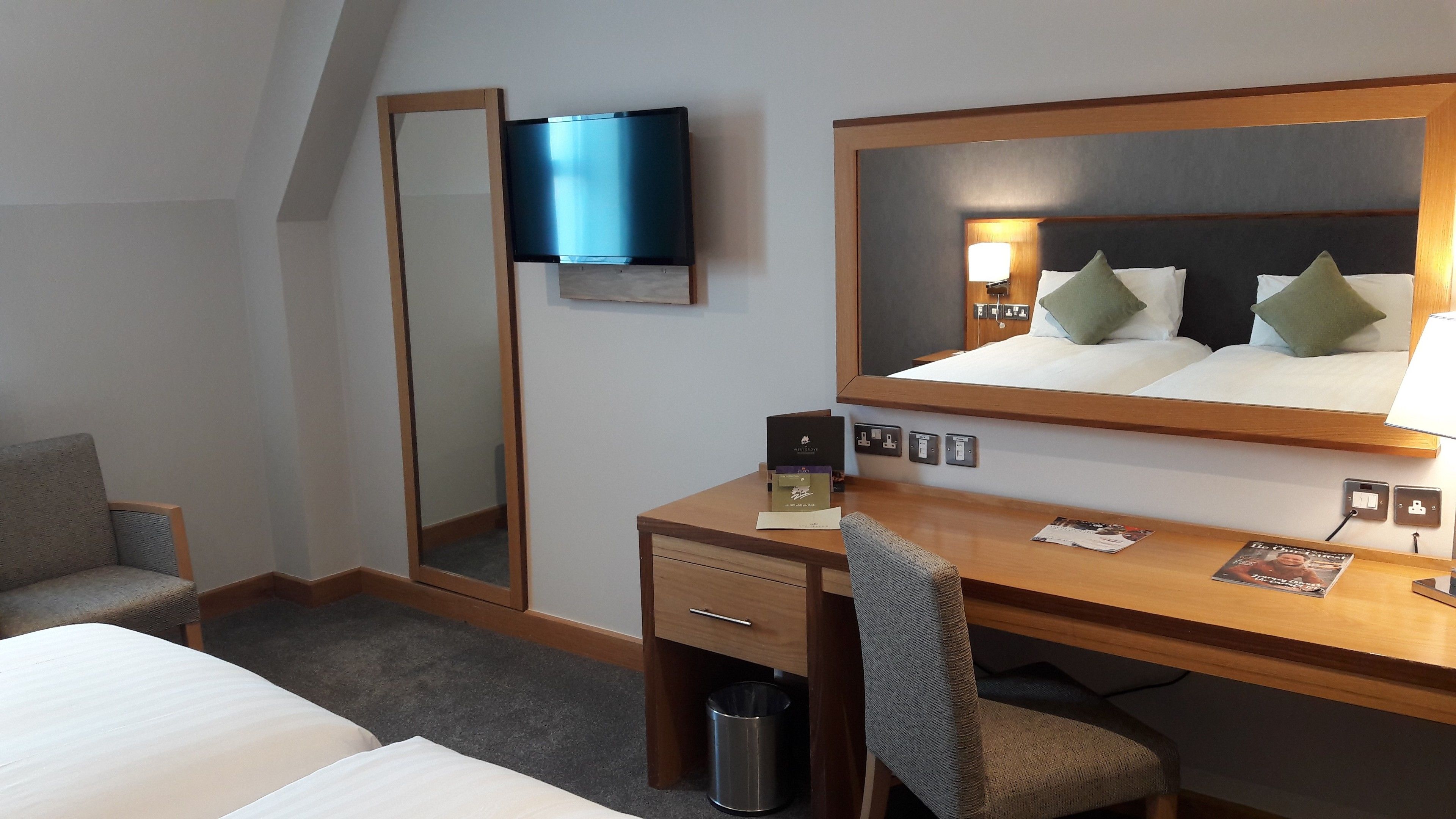 A hotel room with a bed, a desk with a chair, a television, a mirror, and a trash bin.