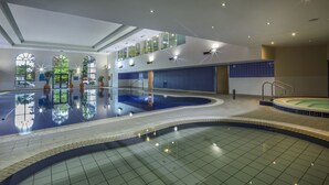 Uma grande piscina interior com água cristalina, rodeada por chão e paredes revestidos de azulejos, e teto com claraboia.