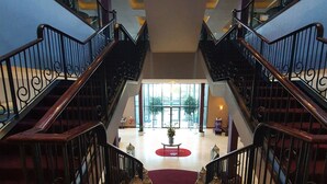 Uma escadaria imponente com grades ornamentadas leva a um amplo hall com tapete vermelho, uma mesa central com um vaso e vista para o exterior através de grandes janelas.