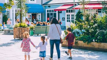 Une femme se promène avec deux enfants sur une place publique, où l’on aperçoit des bancs et, au loin, un magasin Guess.