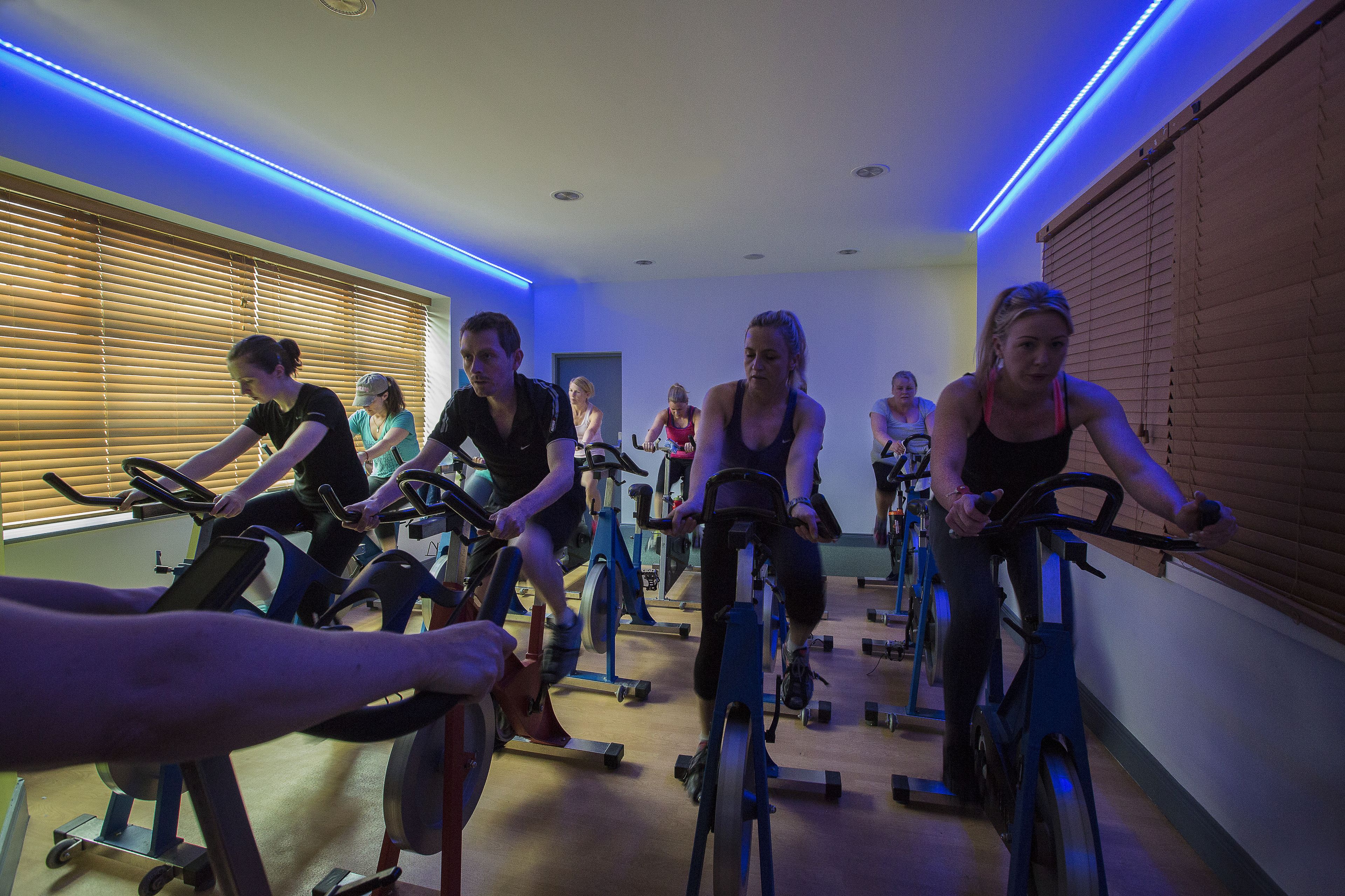 Un grupo de personas haciendo ejercicio en bicicletas estáticas en una habitación con persianas de madera e iluminación LED azul.